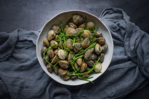 A beautifully arranged bowl of clams served with sprigs of fresh seaweed in a light and minimalist setting. The clams are nestled in a simple, white ceramic bowl with a textured gray cloth as a backdrop. The natural tones and fresh ingredients highlight the refined culinary focus at The Square Club, a popular restaurant in Clifton. This seafood dish reflects the commitment to locally sourced ingredients and sophisticated presentation, making it a signature offering at this highly regarded restaurant in Bristol.