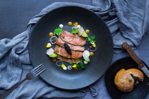 Thinly sliced salmon carpaccio garnished with vibrant edible flowers, black caviar, and a drizzle of sauces on a black plate. This exquisite dish at The Square Club exemplifies its status as one of the finest restaurants in Bristol, perfect for summer dining in the heart of Clifton.