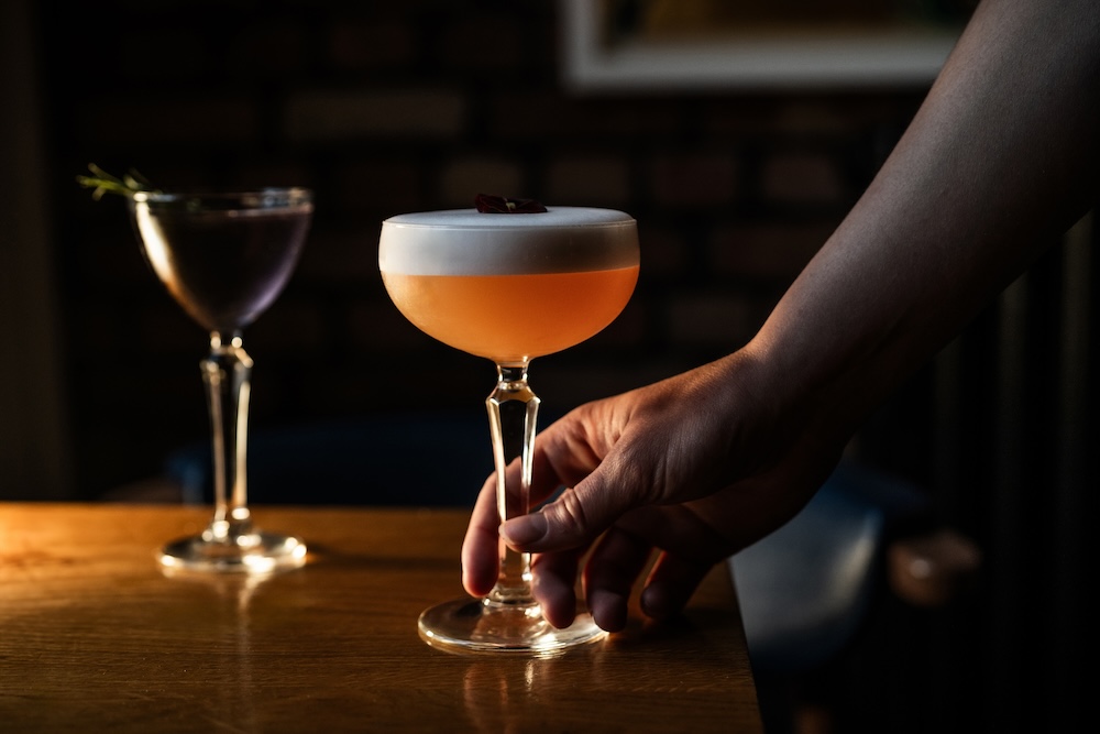 Bartender serving an exquisite signature cocktail with a frothy top, alongside another vibrant drink, at The Square Club restaurant in Bristol. A highlight of the restaurant’s creative cocktail menu in a stylish Clifton setting.